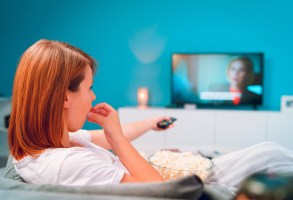 年轻的女人坐在家里的沙发床上，躺在爆米花碗里看电视，盖着毯子，独自一人在她的公寓里享受电影或连续剧，手里拿着遥控器